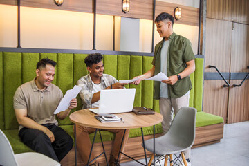 Three Male Coworkers in a Modern Office Discuss Documents While Working on Laptops at Their Desks
