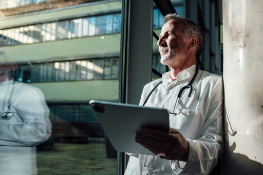 Doctor with stethoscope using tablet looking thoughtfully by window