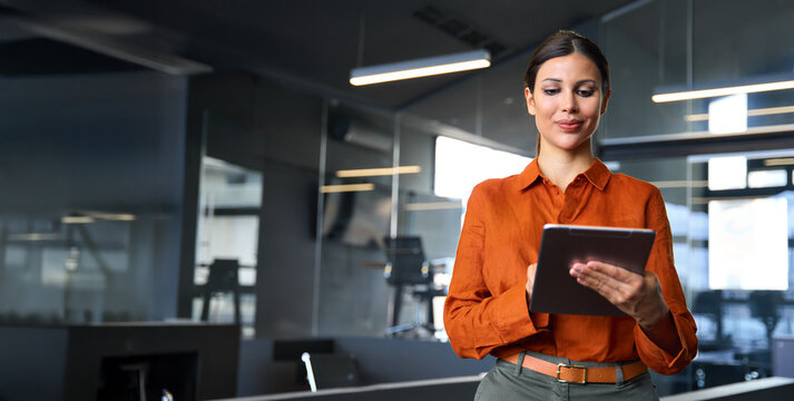 Latin Hispanic mature adult professional business woman looking at online trade app. European businesswoman CEO holding digital tablet using fintech tab application standing at workplace in office.