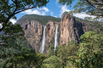 Naklejka premium Waterfall cascading down rocky mountain face, framed by lush foliage