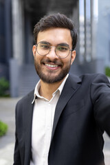 Fototapeta premium Young indian businessman smiling directly at the camera, capturing a self-portrait outdoors in a modern urban environment, showcasing professionalism and a happy demeanor