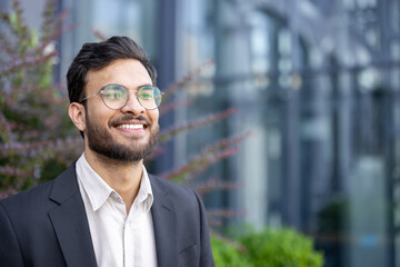Young professional man in a suit jacket and glasses smiling confidently outdoors by an office building, looking toward the future with optimism and ambitious vision for success © Liubomir