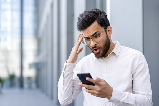 Indian businessman outdoors reacting in shock and worry while reading bad news on his smartphone, showing stress, disbelief and disappointment in a modern urban setting