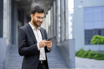 Young businessman wearing glasses and suit standing outdoors against an urban building, smiling while using mobile phone for communication and business