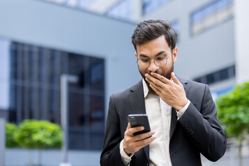 Young businessman experiencing surprise while reading unexpected news on his mobile phone, covering...
