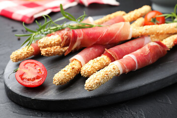 Delicious grissini sticks with sesame seeds, prosciutto and tomato on dark textured table, closeup