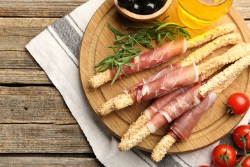Delicious grissini sticks with sesame seeds, prosciutto and other products on wooden table, flat lay. Space for text