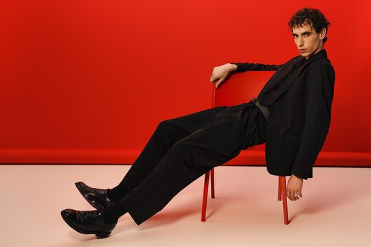 Stylish man exhibits elegance and poise while reclining on a red chair against a bold backdrop