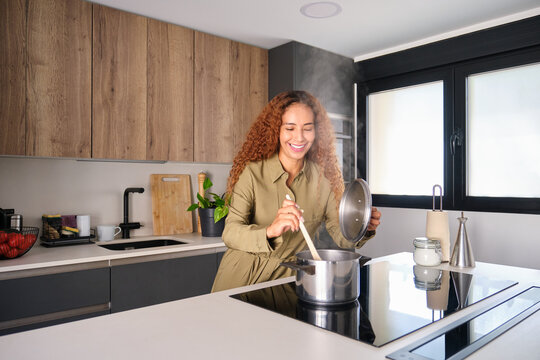 Smiling woman stirring food in pot on induction cooktop in modern kitchen