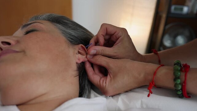Professional therapist carefully inserting a sterile needle into a mature woman's ear during an auricular acupuncture session. Relaxed patient receiving alternative medicine treatment for healing
