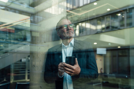 Confident businessman in office holding smartphone and reflecting
