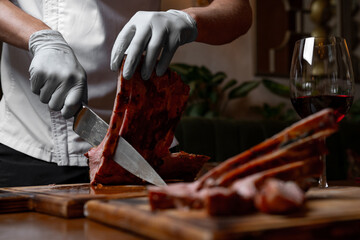 Chef cutting juicy grilled pork ribs on wooden board, close up of hands with knife slicing roasted barbecue meat in restaurant kitchen