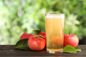 Delicious cider in glass, apples and green leaves on wooden table outdoors