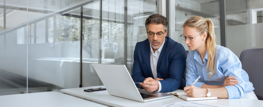 Two partners team of professional business people working discussing writing notes at desk. Mature teacher mentor leader man showing young woman manager project on laptop in office. Banner, copy space