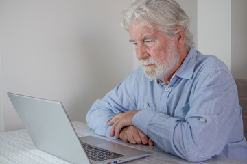 Elderly man looking concentrated while reading on laptop display at his desk, highlighting financial or administrative concerns.