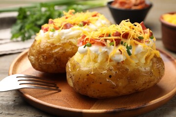 Tasty baked potatoes with sour cream, green onion, cheese, bacon and fork on wooden table, closeup