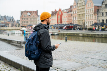 Portrait of a man with a beard in autumn clothes with a smartphone in his hands. Tourist with a backpack in an old European city. Belgium.