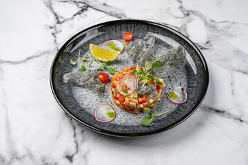 Gourmet salmon tartare with avocado, tomato, microgreens, thin radish slices, black sesame crisps, lemon wedge, and cherry tomato halves served on a modern black plate