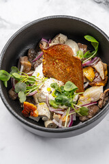Gourmet salad with assorted mushrooms, creamy cheese, red onion, microgreens, and crispy bread served in a black bowl on marble background