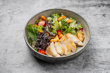 Healthy chicken salad with grilled breast fillet, fresh lettuce, cherry tomatoes, croutons, and creamy dressing served in a ceramic bowl on gray background
