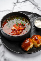 Traditional Ukrainian borscht soup with beetroot, cabbage, pork, fresh herbs, served in black ceramic bowl with sour cream and homemade golden buns on marble background