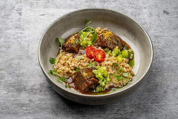 Braised beef with creamy barley risotto, diced cucumber, cherry tomatoes and fresh microgreens served in a ceramic bowl on gray background