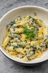 Penne pasta with creamy spinach sauce, grated cheese and fresh herbs served in a ceramic bowl on gray stone background, close up