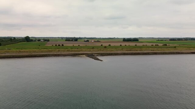 Wide aerial view of a rural landscape with a rocky peninsula jutting into calm water, bordered by farmland, scattered trees, and distant buildings under an overcast grey sky.