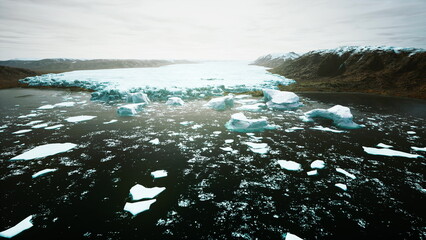 Icy giants break apart and tumble into dark waters, surrounded by rugged mountain peaks under a cloudy sky. A stunning display of natures power and beauty captured in the Arctic. © icetray