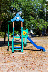 A colorful children's slide with a house-like structure stands in a quiet park, surrounded by trees and sun-drenched ground, evoking a sense of nostalgic innocence.

