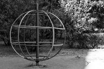 A monochrome image captures a playground spinning orb, its geometric structure standing starkly against the background of trees, evoking a sense of quiet nostalgia.

