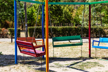 An empty playground swing set with colorful swings and chains, sitting under the sun in a public park with a backdrop of green trees.

