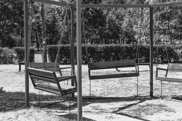 An empty playground swing set in a park on a sunny day, captured in a nostalgic black-and-white style. The swings hang still, waiting for play.

