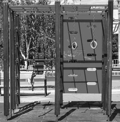A black-and-white image of a modern outdoor playground with a climbing wall, monkey bars, and a hanging rope and rings.

