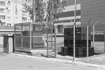 A detailed, black-and-white image of two industrial generators behind security fences with barbed wire, set against an urban backdrop of residential and commercial buildings.

