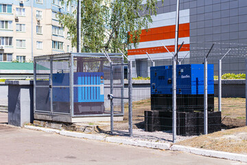 A detailed shot of two large industrial generators, one in a security cage, with barbed wire fencing and a modern building in the background.

