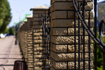 A sunny daytime shot focusing on a sturdy brick fence post with elegant wrought ironwork, extending into a receding line along a paved path with blurred street background.

