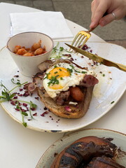 Man slicing breakfast sandwich with fresh bread, cream cheese, crispy bacon, egg, and beans in tomato sauce, cozy cafe meal