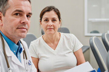 Obraz premium Portrait of man and woman sitting together in medical , man wearing stethoscope, both looking into camera, holding documents