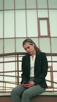 Reflective woman seated in solitude on fence with legs crossed at ankle, staring thoughtfully at phone in hand, dressed in professional outfit, surrounded by modern building and lampposts in urban