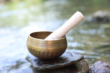 Tibetan singing bowl with mallet on stone near river, closeup