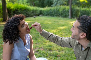 Naklejka premium Interracial couple sharing cherries during romantic picnic in park