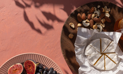 Plate with ripe figs fruit, bunches of grapes, brie cheese and nuts on the mediterranean pink terracotta color plaster background with leaves shadows.