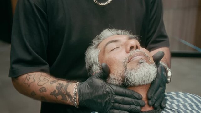 Medium close-up of hands of barber wearing black tshirt, gloves, tattoos massaging aftershave lotion or cologne over neck and face of mature Indian client after shave and beard care treatment