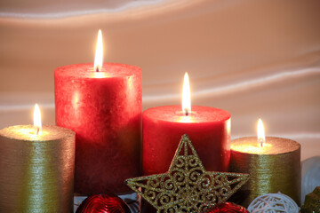 Red and gold burning Christmas candles surrounded by natural potpourri elements and gold stars on a cream satin draped background