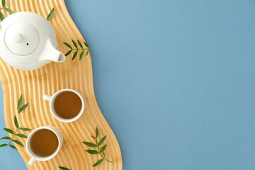 Elegant tea set on wooden tray with green leaves. Minimalist top view against pastel blue.