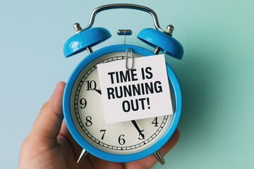 Hand holds blue vintage alarm clock with a stark 'TIME IS RUNNING OUT!' note, symbolizing urgent deadlines and critical business decisions