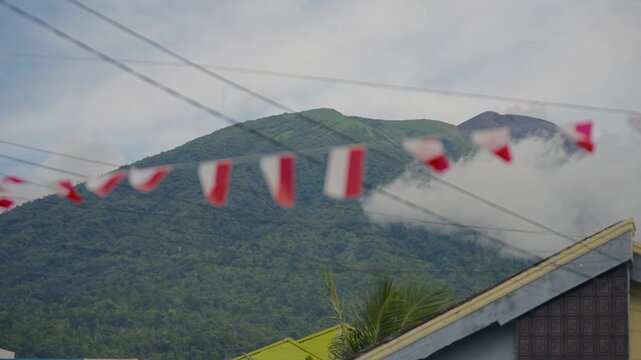 Distant View of a Majestic Green Mountain Peak, Partially Obscured by Clouds, with a Smaller Volcanic Cone Visible in the Background. Blurred red and white flags flutter in the foreground over a build