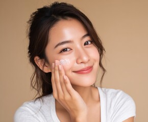 Fototapeta premium A young woman applies facial cream to her cheek, showcasing healthy skin and a serene expression against a soft beige background.