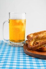 Beer mug and pretzel loaf on a wooden tray with a blue checkered tablecloth or napkin on a white table. Concept of Oktoberfest. Bavarian German culture festival decoration symbol